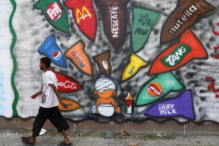 A man walks past a mural in which consumer brand logos are painted as missiles aimed at a bandaged baby holding a Palestinian flag